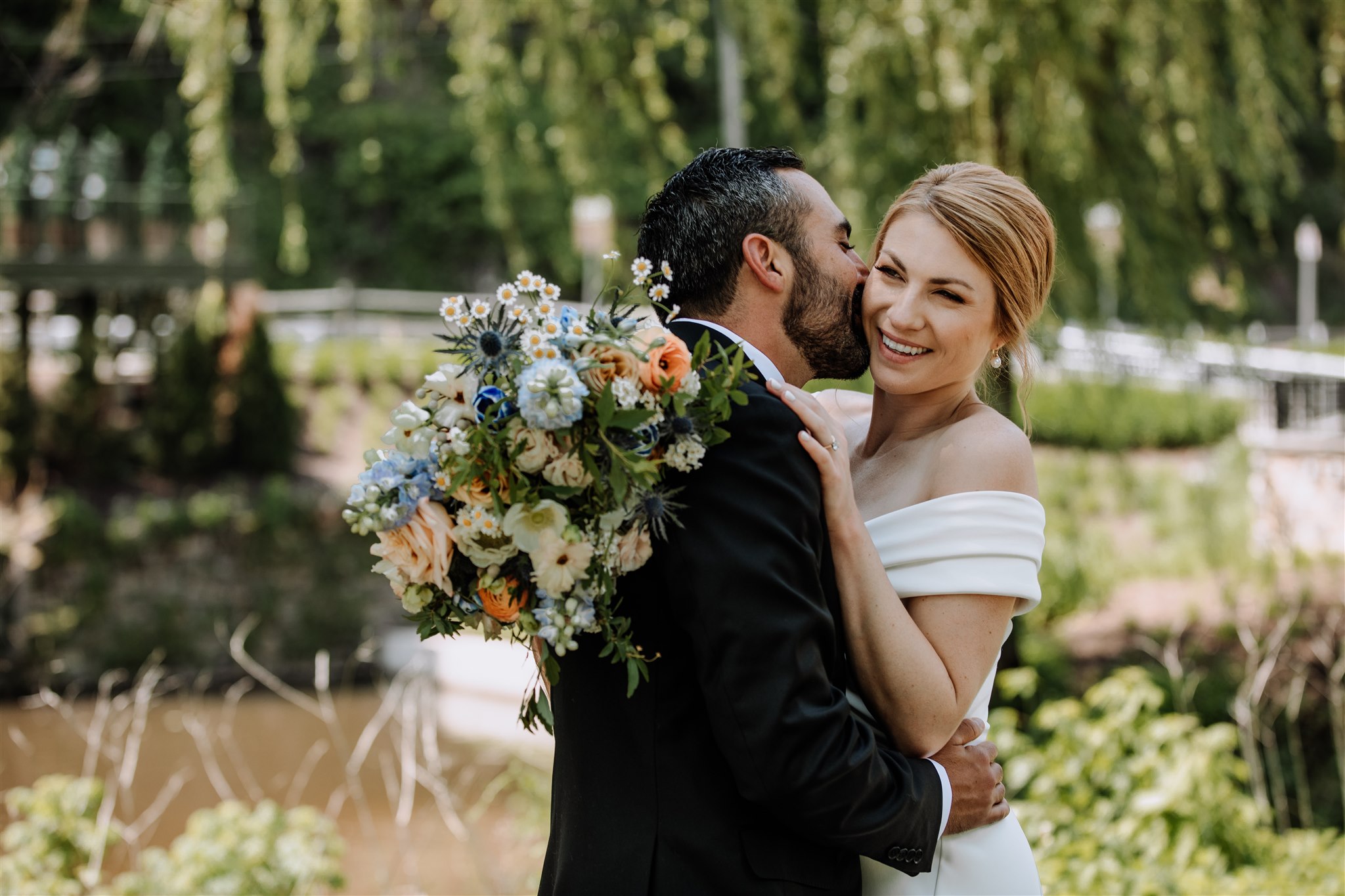 Groom kissing his smiling bride while she holds a bouquet of flowers with green trees behind them