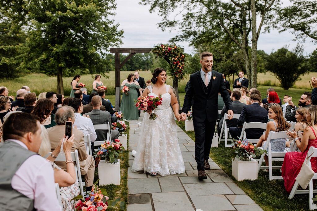 The Farm Bakery & Events in PA with a recently married bride and groom walking while wearing a white dress and blue suit with florals and guests behind them.